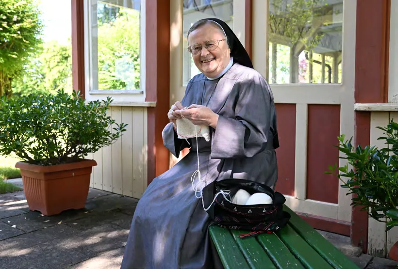 Eine Schwester sitzt strickend auf einer Bank vor dem Gartenhaus